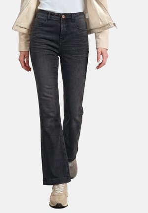 Persoon die zwarte bootcut jeans met hoge taille, beige jas, witte geribbelde top en beige sneakers draagt en vooruit loopt tegen een witte achtergrond.