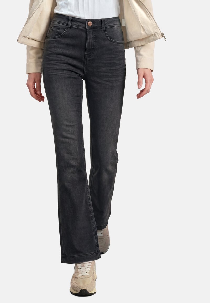 Personne portant un jean noir bootcut taille haute, une veste beige, un top blanc côtelé et des baskets beiges, marchant vers l'avant sur fond blanc.