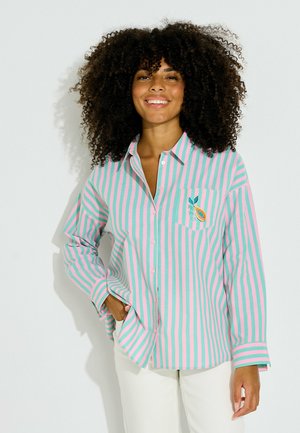 Mujer con cabello rizado que lleva una camisa de rayas rosas y verdes con botones y un parche de fruta, sonriendo con una mano en el bolsillo, pantalones blancos.