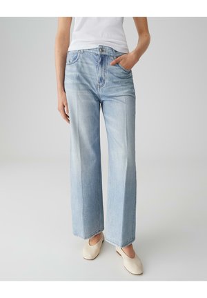 Persoon draagt lichtblauwe wijd uitlopende spijkerbroek en witte instapschoenen, met één hand in de zak en een wit shirt.