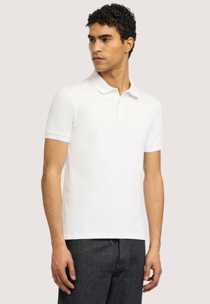 Mann trägt ein schlichtes weißes Poloshirt mit kurzen Ärmeln und dunkle Jeans, steht und schaut seitlich vor einem einfarbigen hellen Hintergrund.