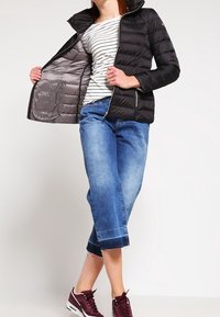 Doudoune noire avec fermeture éclair à l'avant, texture matelassée et doublure grise. Port ée par-dessus un haut rayé, associée à un jean bleu court et des baskets bordeaux.