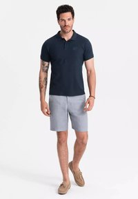 Polo de color marino con mangas cortas y collar, combinado con pantalones cortos grises claros y zapatos cómodos marrones. Presenta un logotipo sutil en el pecho.