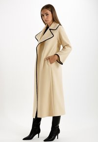 Donna in un lungo cappotto beige con dettagli neri, mani nelle tasche, in piedi e con stivali neri con tacco su sfondo bianco.