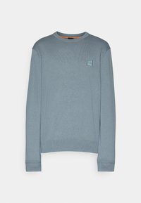 Pull bleu clair à col ras-du-cou avec manches longues et petite écusson carré logo sur la poitrine gauche sur un fond uni.