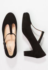 Zapatos de tacón de ante negro con un diseño de tira en T, lados abiertos y un tacón ancho. El interior tiene un forro suave en color beige con el logotipo de la marca.