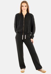 Sudadera negra con cremallera, mangas largas y bolsillos laterales, combinada con pantalones deportivos negros con cordón. Textura de tela suave.