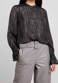 Blusa nera in metallo con maniche lunghe, superficie texturizzata e colletto alto, abbinata a pantaloni in pelle grigi chiari con cintura.