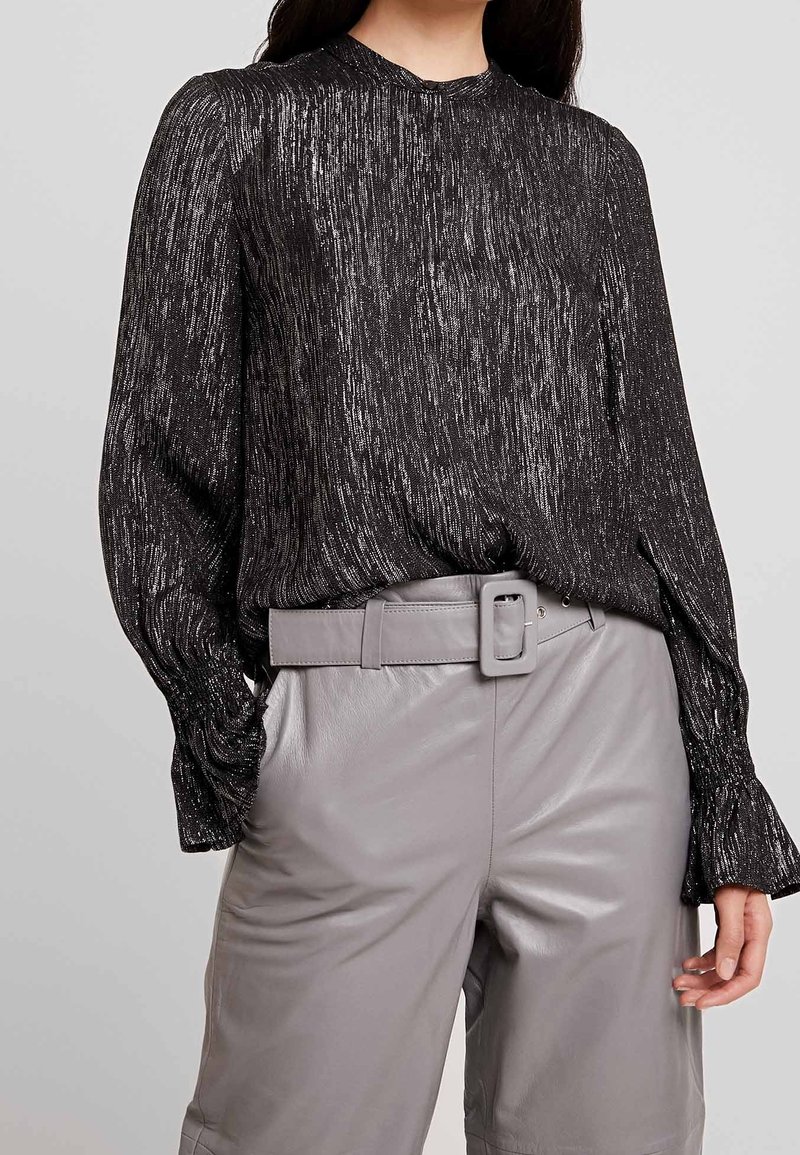 Blusa nera in metallo con maniche lunghe, superficie texturizzata e colletto alto, abbinata a pantaloni in pelle grigi chiari con cintura.