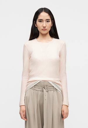 Světle růžová košile s dlouhým rukávem a kulatým výstřihem, vyrobená z měkké tkaniny. Kombinována s béžovými kalhotami s vysokým pasem, vybavenými páskem a sklady.