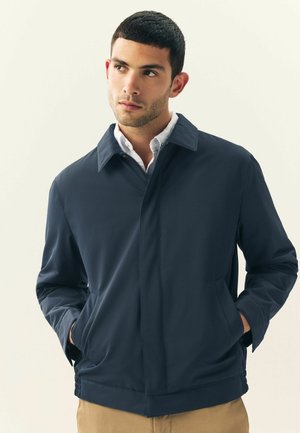 Chaqueta ligera de color azul marino con cierre de cremallera, cuello puntiagudo y bolsillos laterales; presenta una textura suave y un diseño minimalista.