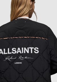 Czarna, pikowana kurtka o matowym wykończeniu, z kontrastującym lamowaniem w panterkę i dużym, białym napisem "ALLSAINTS" na plecach.