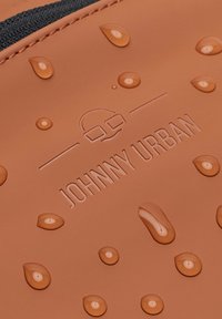 Vanddråber på en glat, tanfarvet overflade præget med "Johnny Urban"-logoet, der har et lille ikon over teksten.