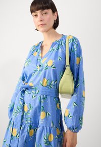 Rochie albastră cu imprimeu de lămâi și flori, cu mâneci lungi și curea la talie. Complementează o geantă verde texturată, mică, cu curea de umăr.
