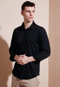 Zwart overhemd met lange mouwen en knoopsluiting, met kraag en een gladde textuur, gedragen door een model in een neutrale setting.