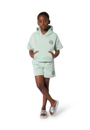 Young girl with hands in pockets wears mint green hooded short-sleeve top, matching shorts, and silver slides, standing with crossed legs.