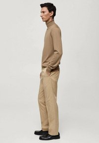 Maglione beige a collo alto abbinato a pantaloni beige. L'insieme presenta una vestibilità rilassata e una consistenza liscia, completato da scarpe nere.