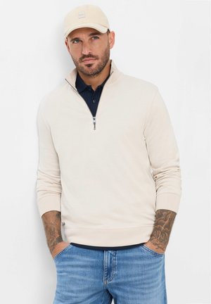 Mann trägt beigen Pullover mit Viertel-Reißverschluss, marineblaues Poloshirt, beichen Cap und blaue Jeans, steht mit Händen in den Taschen vor weißem Hintergrund.