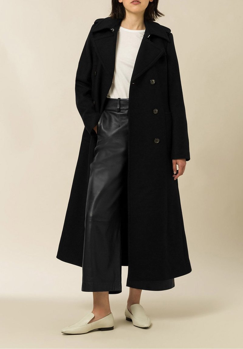 Femme portant un long manteau noir, une chemise blanche, un pantalon en cuir noir à jambes larges et des mocassins blancs, debout devant un fond uni.