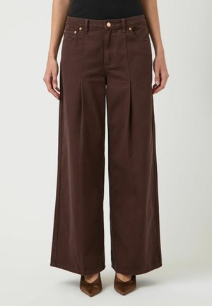 Kvinde iført brune bukser med høj talje og vide ben med forreste folder og kobberknapper, kombineret med brune spidse sko.