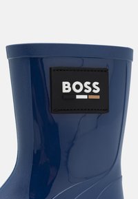 Botte de pluie en caoutchouc bleu marine avec une surface texturée et un patch logo "BOSS" noir et blanc très visible sur le côté extérieur.