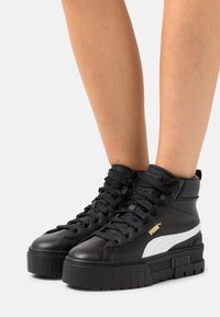 Svarta högstövlar från Puma med vit rand och tjock sula, på bar hud mot en enkel ljus bakgrund.