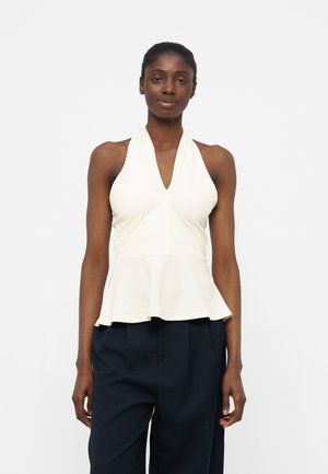 Γυναίκα που φορά λευκό αμάνικο τοπ με V λαιμόκοψη και βολάν στη μέση, σε συνδυασμό με σκούρο ψηλόμεσο παντελόνι, όρθια μπροστά σε λευκό φόντο.