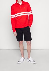 Rød langærmet polo med hvide striber, med en knappet krave og gyldent logo. Parret med sorte shorts og hvidternede sneakers.