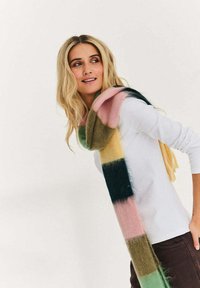 Blondynka ubrana w białą koszulę z długim rękawem i kolorowy, paskowany szalik pochyla się do przodu, patrząc w bok z lekkim uśmiechem.