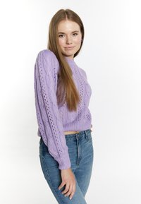 Lavendelfarbener Strickpullover mit Kabelmustern, langen Ärmeln mit gerippten Bündchen, kombiniert mit hellblauen hochgeschnittenen Jeans.
