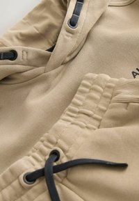 Detailný záber na béžovú mikinu s kapucňou a tepláky s čiernymi šnúrkami a elastickým pásom, zobrazujúci textúru látky a detaily šitia.