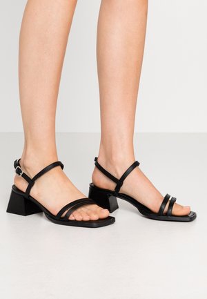 Black strappy heeled sandals with a square toe, featuring two thin straps across the foot and a block heel, made from smooth leather.
