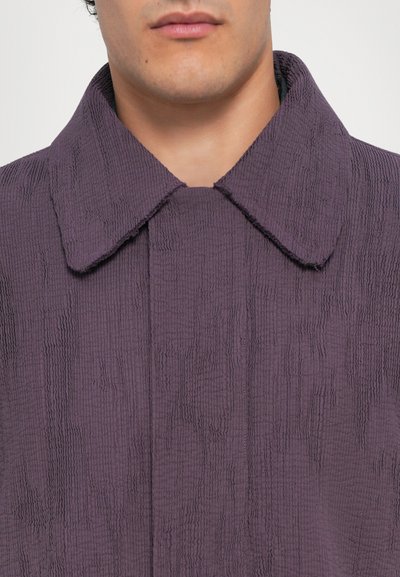 Lila strukturierte Jacke mit einem ausgeprägten Kragen, versehen mit wellenförmigen Mustern und einem offenen Halsausschnitt, aus weichem Material gefertigt.