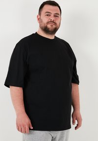 Černé tričko s krátkým rukávem vyrobené z bavlny. Jednoduchý, kruhový výstřih. Volný střih rovného střihu. Nosené s šedými tepláky.