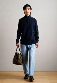 Marineblå lynlås striktrøje, lyseblå lige bukser, brune slip-on sko og grøn taske med korte håndtag. Trægulv i baggrunden.