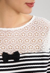 Valkoinen pitsipaita, jossa on kukkakuosi, musta raidallinen design ja pieni musta nauha keskellä.