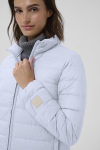 Helle blaue Puffjacke mit horizontalen Nähten, hohem Kragen und einem strukturierten grauen Rollkragenpullover darunter. Auffälliges Etikett am Ärmel.