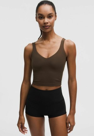 Donna che indossa un top corto marrone senza maniche e pantaloncini neri a vita alta, in piedi contro uno sfondo chiaro e uniforme.