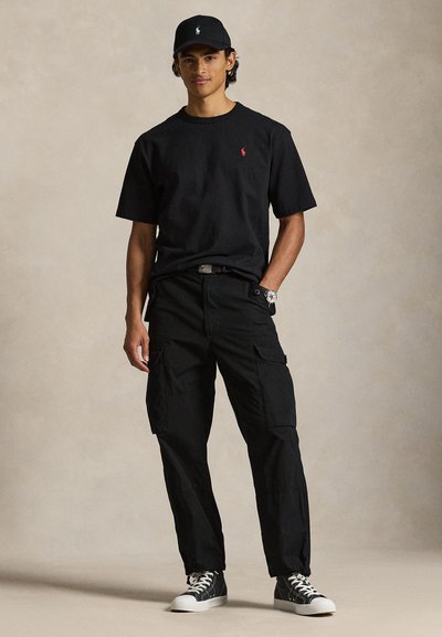 Μαύρο βαμβακερό μπλουζάκι με μικρό κόκκινο λογότυπο, σε συνδυασμό με μαύρο cargo παντελόνι με πλαϊνές τσέπες και μαύρα και λευκά αθλητικά παπούτσια. Μαύρο καπέλο.