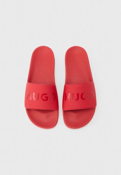 Paire de sandales slides rouges avec semelles texturées et larges brides embossées du nom de la marque "HUGO" sur un fond neutre.