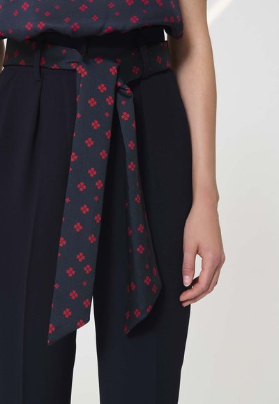Pantalon bleu marine avec une ceinture en tissu assorti nouée en un nœud, présentant un motif floral rouge, porté avec un haut assorti à motif similaire.