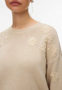 Pull en tricot beige avec des motifs floraux en relief sur l'épaule et le haut de la poitrine, porté par une personne avec une boucle d'oreille chaîne pendante.