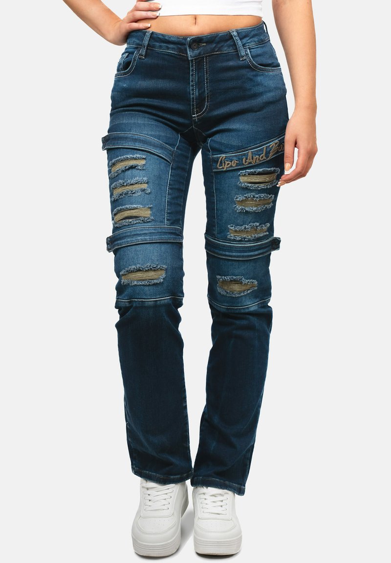 Jeans in denim blu scuro con dettagli strappati, inclusi buchi e toppe. Design aderente con chiusura a bottone e cerniera. Tasche posteriori semplici.