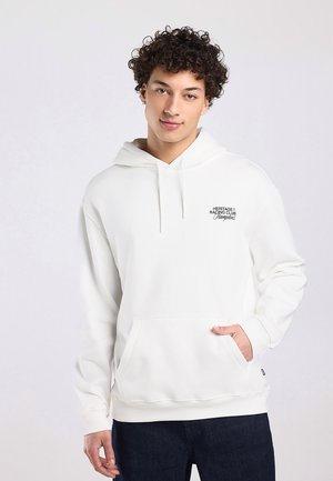 Giovane con capelli ricci che indossa una felpa bianca con cappuccio "Heritage Racing Club Hamptons" e jeans scuri, con una mano nella tasca frontale.