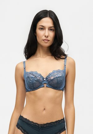 Femme aux longs cheveux foncés portant un soutien-gorge et une culotte en dentelle bleue assortie sur un fond clair uni.
