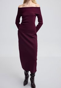 Robe bordeaux à épaules dénudées avec manches longues, design ajusté et jupe superposée. Associée à des bottes noires à finition brillante.