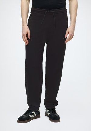 Osoba odjevena u crne trenirke s vezicama uparene s crno-bijelim tenisicama, stoji na jednobijeloj pozadini.