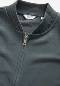 Sweat à capuche zippé gris foncé, avec col et poignets côtelés, texture lisse, et une fermeture éclair en métal argenté avec une tirette carrée.