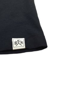 Schwarzes Baumwoll-T-Shirt mit Saum, der ein gewebtes weißes Etikett mit schwarzem Text und kreuzenden Designelementen zeigt. Glatte Textur und simples Design.