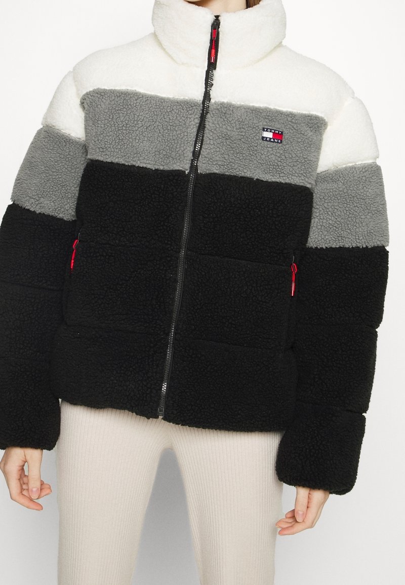 Giacca in pile con colletto alto, caratterizzata da blocchi di colore nero, grigio e bianco. Chiusura con zip e patch con logo ben visibile sul petto.